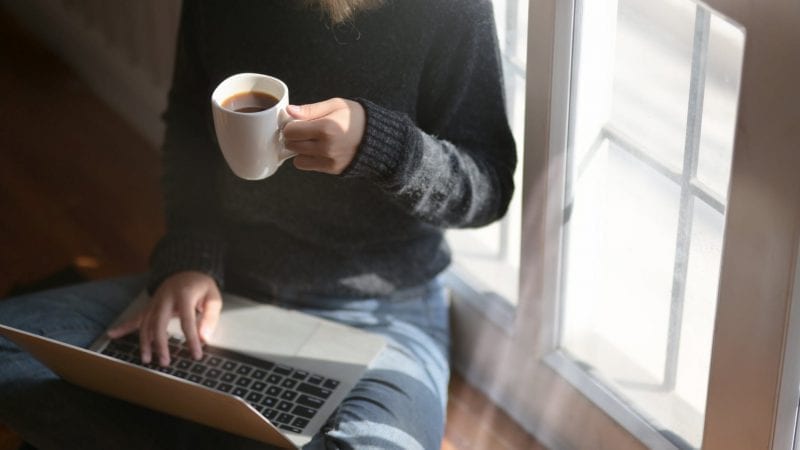 Een vrouw die multitaskt met een kopje koffie en een laptop terwijl ze op afstand werkt.